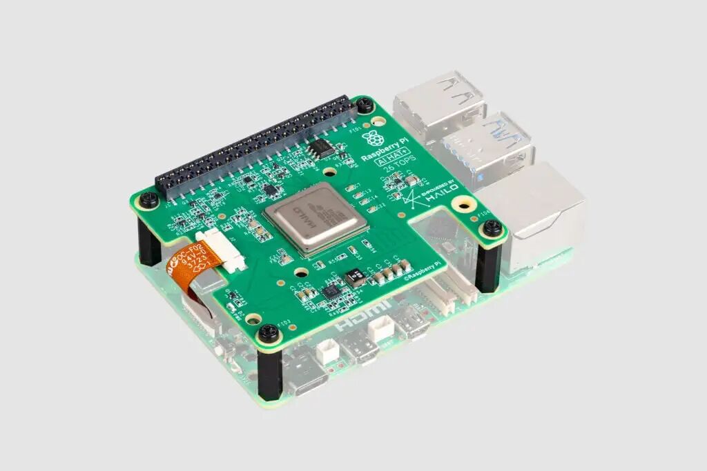 Raspberry Pi single-board computer with an attached AI accelerator module, likely the Raspberry Pi AI Hat. This hat includes a green circuit board with a central chip that appears to be from Hailo, a company that specializes in artificial intelligence (AI) processors. The board is connected to the Raspberry Pi via the GPIO pins, and it has several components related to AI processing and other features to enable high-performance machine learning on the device. This configuration is designed for AI applications like real-time image processing, neural network acceleration, and other computationally intensive tasks. The text "26 TOPS" refers to the AI hat's ability to perform 26 trillion operations per second, which is a significant performance specification for AI applications.